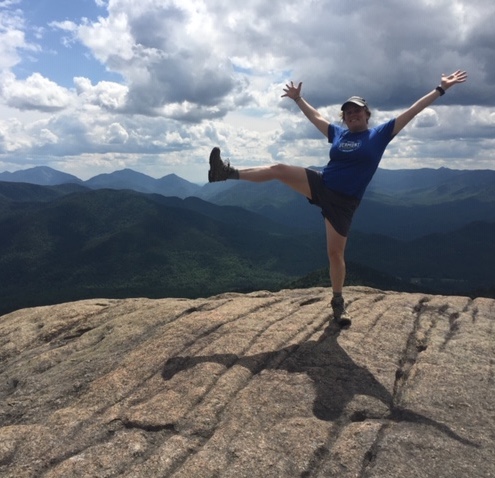 Carina smiling and standing on one leg on a mountaintop with a landscape behind her.
