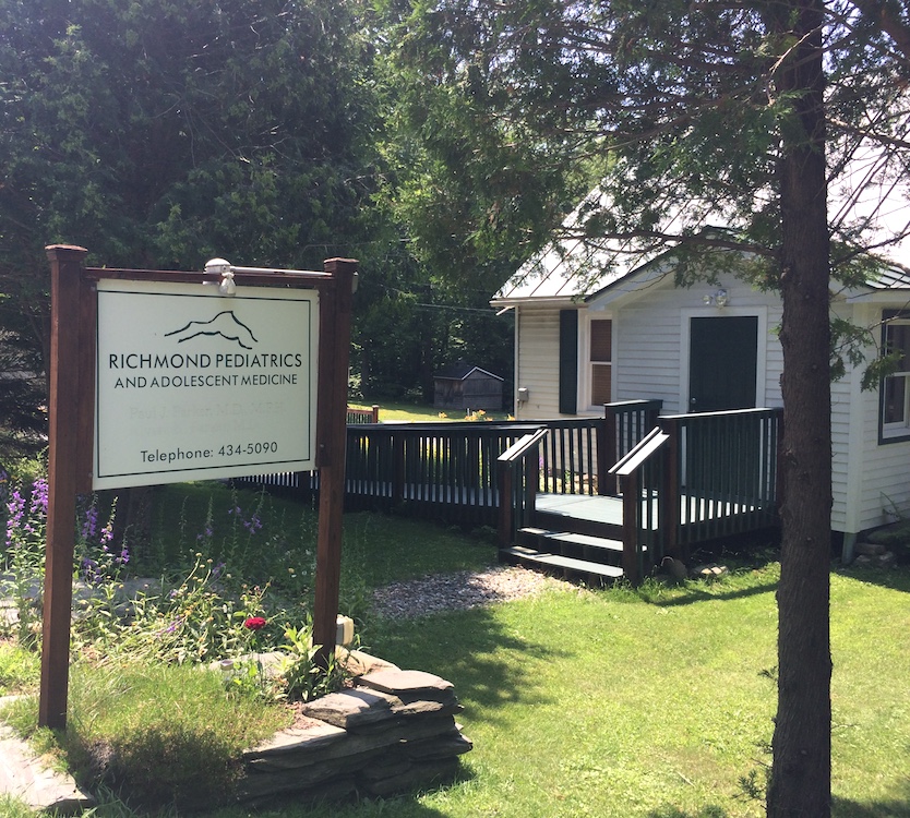 The sign and front door of the Richmond Pediatrics office. There are flowers underneath the sign. There are stairs and a ramp that lead up to the front door of the office.